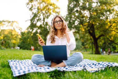 Neşeli kadın güneşli bir parkta dizüstü bilgisayarla battaniyeye oturur ve kablosuz kulaklıkla müzik dinler. Dışarıda telefonu ve dizüstü bilgisayarı olan güzel bir kadın çalışıyor. Serbest konsept, teknoloji.