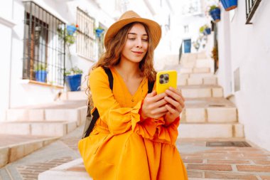 Şapkalı ve telefonlu parlak elbiseli genç bir turist güzel bir kasabanın merdivenlerinde oturuyor. Güzel bir kadın güneşli havanın tadını çıkarır. Turizm, blog konsepti.