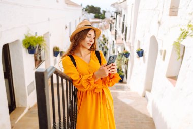 Şapkalı ve telefonlu parlak elbiseli genç bir turist güzel bir kasabanın merdivenlerinde oturuyor. Güzel bir kadın güneşli havanın tadını çıkarır. Turizm, blog konsepti.