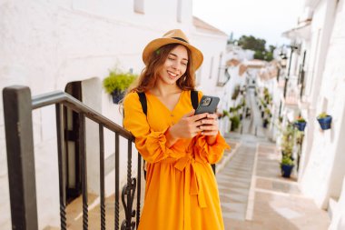 Şapkalı ve telefonlu parlak elbiseli genç bir turist güzel bir kasabanın merdivenlerinde oturuyor. Güzel bir kadın güneşli havanın tadını çıkarır. Turizm, blog konsepti.