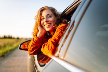 Mutlu kadın arabanın camından sarkıyor ve pencereden manzaranın tadını çıkarıyor. Genç bayan gezgin yol macerasında özgürlüğü hissediyor. Seyahat, özgürlük konsepti. Etkin yaşam biçimi.