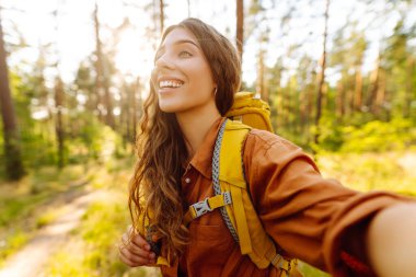 Parlak sarı sırt çantalı genç bayan yürüyüşçünün portresi sık ormanda selfie çekerken görülüyor. Güzel bir kadın seyahat ediyor, doğanın tadını çıkarıyor. Macera, doğa konsepti.