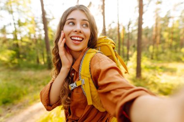 Parlak sarı sırt çantalı genç bayan yürüyüşçünün portresi sık ormanda selfie çekerken görülüyor. Güzel bir kadın seyahat ediyor, doğanın tadını çıkarıyor. Macera, doğa konsepti.