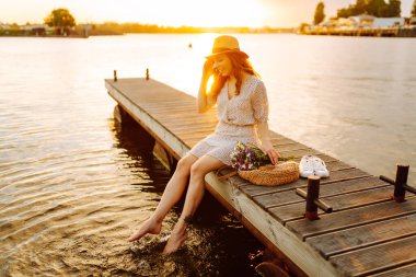 Şapkalı neşeli bir kadın ve elinde bir buket kır çiçeğiyle iskelede gün batımının tadını çıkarıyor. Tatlı bir kadın gün batımıyla karşılaşır ve güneşin ışınlarını yakalar. Rahatlama konsepti, güzellik.