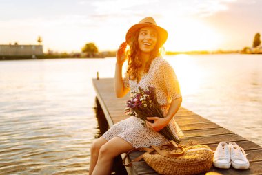 Şapkalı neşeli bir kadın ve elinde bir buket kır çiçeğiyle iskelede gün batımının tadını çıkarıyor. Tatlı bir kadın gün batımıyla karşılaşır ve güneşin ışınlarını yakalar. Rahatlama konsepti, güzellik.