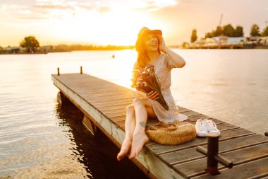 Şapkalı neşeli bir kadın ve elinde bir buket kır çiçeğiyle iskelede gün batımının tadını çıkarıyor. Tatlı bir kadın gün batımıyla karşılaşır ve güneşin ışınlarını yakalar. Rahatlama konsepti, güzellik.