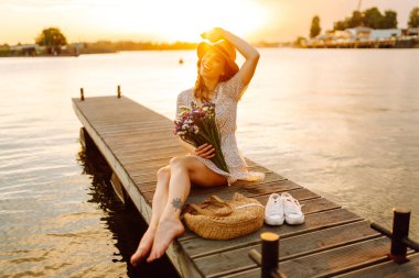 Şapkalı neşeli bir kadın ve elinde bir buket kır çiçeğiyle iskelede gün batımının tadını çıkarıyor. Tatlı bir kadın gün batımıyla karşılaşır ve güneşin ışınlarını yakalar. Rahatlama konsepti, güzellik.