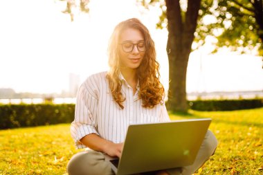 Parktaki yeşil çimlerin üzerinde dizüstü bilgisayarı olan gülümseyen kadın. Parkta çalışan ve gün batımının tadını çıkaran güzel bir kadın serbest yazar. Doğa, eğlence konsepti.