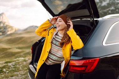 Parlak yağmurluklu gezgin araba bagajında oturur ve dağ manzarasının tadını çıkarır. Arabaya yakın güzel bir kadın yolda özgürlüğü hissediyor. Doğa, seyahat konsepti, yolculuk.