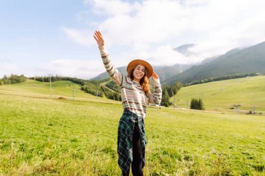 Genç bayan gezgin yeşil tepelik çayırda dağ manzarasının tadını çıkarıyor. Dışarıda özgürlüğünü hisseden şapkalı güzel bir kadın. Seyahat, doğa, yürüyüş konsepti.