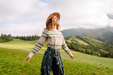 Genç bayan gezgin yeşil tepelik çayırda dağ manzarasının tadını çıkarıyor. Dışarıda özgürlüğünü hisseden şapkalı güzel bir kadın. Seyahat, doğa, yürüyüş konsepti.