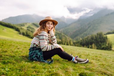 Genç bayan gezgin yeşil tepelik çayırda dağ manzarasının tadını çıkarıyor. Dışarıda özgürlüğünü hisseden şapkalı güzel bir kadın. Seyahat, doğa, yürüyüş konsepti.