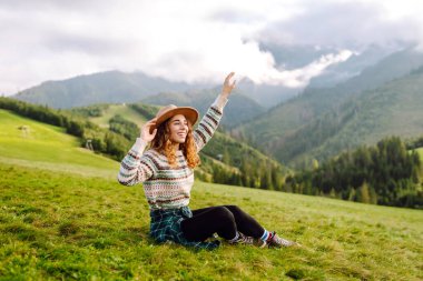 Genç bayan gezgin yeşil tepelik çayırda dağ manzarasının tadını çıkarıyor. Dışarıda özgürlüğünü hisseden şapkalı güzel bir kadın. Seyahat, doğa, yürüyüş konsepti.