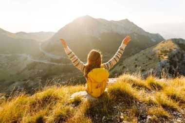 Dağ zirvesinde duran parlak sırt çantalı mutlu kadın yürüyüşçünün arka görüntüsü. Genç bir kadın dağ manzarasının tadını çıkarıyor ve gün batımında özgürlüğü hissediyor. Yürüyüş, spor konsepti. Etkin yaşam biçimi.