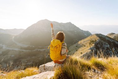 Dağ zirvesinde duran parlak sırt çantalı mutlu kadın yürüyüşçünün arka görüntüsü. Genç bir kadın dağ manzarasının tadını çıkarıyor ve gün batımında özgürlüğü hissediyor. Yürüyüş, spor konsepti. Etkin yaşam biçimi.