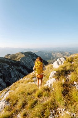 Dağ zirvesinde duran parlak sırt çantalı mutlu kadın yürüyüşçünün arka görüntüsü. Genç bir kadın dağ manzarasının tadını çıkarıyor ve gün batımında özgürlüğü hissediyor. Yürüyüş, spor konsepti. Etkin yaşam biçimi.