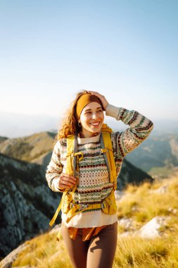 Dağ zirvesinde duran parlak sırt çantalı mutlu kadın yürüyüşçünün arka görüntüsü. Genç bir kadın dağ manzarasının tadını çıkarıyor ve gün batımında özgürlüğü hissediyor. Yürüyüş, spor konsepti. Etkin yaşam biçimi.