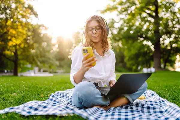 Neşeli kadın güneşli bir parkta dizüstü bilgisayarla battaniyeye oturur ve kablosuz kulaklıkla müzik dinler. Dışarıda telefonu ve dizüstü bilgisayarı olan güzel bir kadın çalışıyor. Serbest konsept, teknoloji.