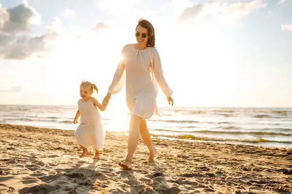Kumsalda kızıyla birlikte gün batımında birlikte eğlenen mutlu anne portresi. Genç bir kadın ve küçük bir kız gülüyor ve deniz manzarasının tadını çıkarıyor. Çocukluk, annelik kavramı..