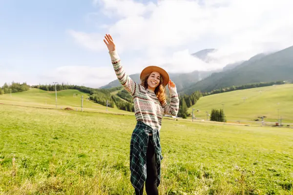 Genç bayan gezgin yeşil tepelik çayırda dağ manzarasının tadını çıkarıyor. Dışarıda özgürlüğünü hisseden şapkalı güzel bir kadın. Seyahat, doğa, yürüyüş konsepti.