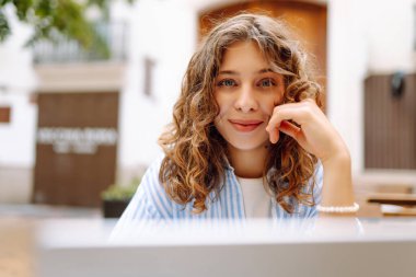 Bilgisayarlı mutlu kadın masada çalışıyor ve terasta kahve içiyor. Genç bayan serbest yazar dışarıda kahve içiyor. Uzak çalışma konsepti, içkiler, rahatlama.