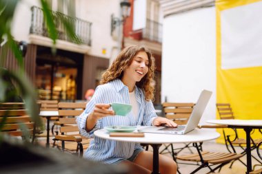 Bilgisayarlı mutlu kadın masada çalışıyor ve terasta kahve içiyor. Genç bayan serbest yazar dışarıda kahve içiyor. Uzak çalışma konsepti, içkiler, rahatlama.