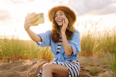 Selfie zamanı! Kumsalda telefonu olan mutlu bir kadın sahilde günbatımının tadını çıkarıyor. Şapkalı ve çantalı genç bir turist doğanın tadını çıkarıyor. Bloglama ve teknoloji kavramı.