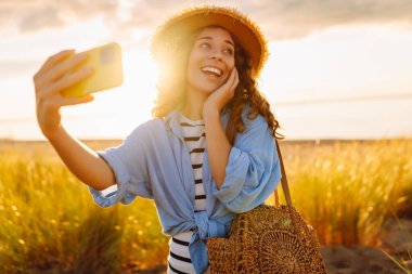 Selfie zamanı! Kumsalda telefonu olan mutlu bir kadın sahilde günbatımının tadını çıkarıyor. Şapkalı ve çantalı genç bir turist doğanın tadını çıkarıyor. Bloglama ve teknoloji kavramı.
