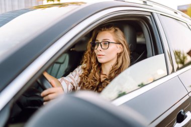 Gözlüklü genç kadın sürücü arabasından dışarı bakıyor. Sürücü koltuğundaki güzel kadın güneşli bir günün tadını çıkarıyor. Ulaşım, modernlik ve eğlence kavramı.