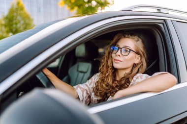 Gözlüklü genç kadın sürücü arabasından dışarı bakıyor. Sürücü koltuğundaki güzel kadın güneşli bir günün tadını çıkarıyor. Ulaşım, modernlik ve eğlence kavramı.