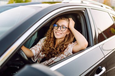 Gözlüklü genç kadın sürücü arabasından dışarı bakıyor. Sürücü koltuğundaki güzel kadın güneşli bir günün tadını çıkarıyor. Ulaşım, modernlik ve eğlence kavramı.