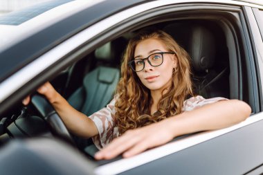 Gözlüklü genç kadın sürücü arabasından dışarı bakıyor. Sürücü koltuğundaki güzel kadın güneşli bir günün tadını çıkarıyor. Ulaşım, modernlik ve eğlence kavramı.