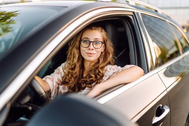 Gözlüklü genç kadın sürücü arabasından dışarı bakıyor. Sürücü koltuğundaki güzel kadın güneşli bir günün tadını çıkarıyor. Ulaşım, modernlik ve eğlence kavramı.