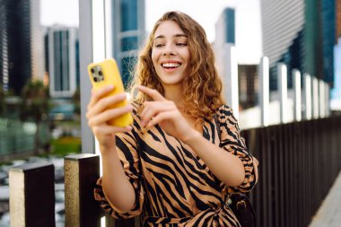 Ellerinde telefon olan gülümseyen bir kadın şehrin telaşına karşı. Genç bir kadın dışarıda telefonuyla blog yazıyor ve sohbet ediyor. Blogculuk, teknoloji, turizm kavramı.