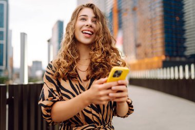 Ellerinde telefon olan gülümseyen bir kadın şehrin telaşına karşı. Genç bir kadın dışarıda telefonuyla blog yazıyor ve sohbet ediyor. Blogculuk, teknoloji, turizm kavramı.