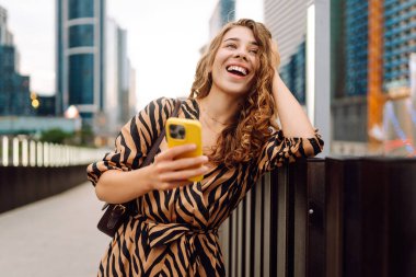 Ellerinde telefon olan gülümseyen bir kadın şehrin telaşına karşı. Genç bir kadın dışarıda telefonuyla blog yazıyor ve sohbet ediyor. Blogculuk, teknoloji, turizm kavramı.