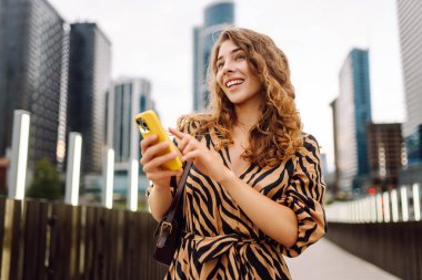 Ellerinde telefon olan gülümseyen bir kadın şehrin telaşına karşı. Genç bir kadın dışarıda telefonuyla blog yazıyor ve sohbet ediyor. Blogculuk, teknoloji, turizm kavramı.