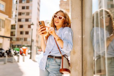 Şehir yürüyüşünden zevk alan genç bir kadının portresi. Elinde telefonla. Güzel bir kadın blog yazıyor, gülümsüyor ve açık havada güneşli havanın tadını çıkarıyor. Turizm ve yürüyüş konsepti. Yaşam biçimi.