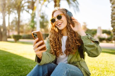 Güneş gözlüğü takan, elinde telefon tutan ve güneşli bir parkta eğlenen genç bir kadın. Müzik dinleyen güzel bir turist ya da dışarıda kablosuz kulaklıklardan yayın yapan güzel bir turist. Yürüyen, rahatlatıcı konsept.