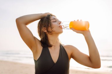 Spor giyimli güzel bir kadın deniz manzarasına karşı renkli bir şişeden su içiyor. Sporcu bir kadın antrenmandan zevk alıyor ve doğadaki susuzluğunu gideriyor. Spor ve rahatlama konsepti.