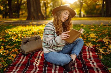 Güzel bir kadının portresi, elinde bir kitapla güneşli bir ormanda battaniyede dinleniyor. Açık havada kitap okumaktan zevk alan gülümseyen bir kadın. Rahatlama, doğa ve sonbahar konsepti.