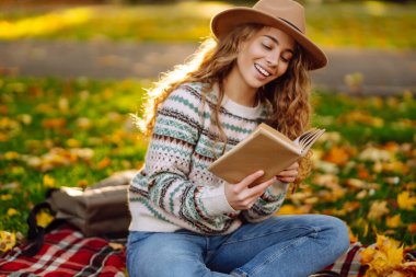 Güzel bir kadının portresi, elinde bir kitapla güneşli bir ormanda battaniyede dinleniyor. Açık havada kitap okumaktan zevk alan gülümseyen bir kadın. Rahatlama, doğa ve sonbahar konsepti.