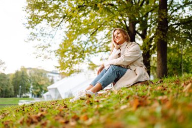 Genç bir kadın nehrin yakınındaki bir sonbahar parkında bir battaniyenin üzerinde dinleniyor. Neşeli kadın açık havada güneşli havayı seviyor, yeşil çimlerde oturuyor. Yaşam tarzı. Güzellik, rahatlama konsepti.