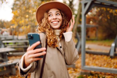 Şapkalı ve telefonlu genç bir kadın sonbahar parkındaki masada oturuyor. Güzel kadın blogları ve açık havada güneşli bir günün tadını çıkarıyor. Bloglama, yürüme ve eğlenme konsepti.