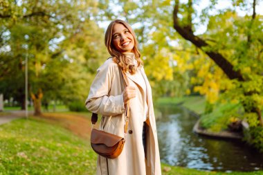Gündelik giysiler içinde güzel bir kadın sonbahar parkında yürüyor ve güneşli havanın tadını çıkarıyor. Açık havada poz veren hoş bir kadın. Güzellik ve hafta sonu konsepti. Yaşam biçimi.