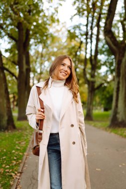 Gündelik giysiler içinde güzel bir kadın sonbahar parkında yürüyor ve güneşli havanın tadını çıkarıyor. Açık havada poz veren hoş bir kadın. Güzellik ve hafta sonu konsepti. Yaşam biçimi.