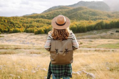 Kayalık bir arazide duran sırt çantalı bir kadın yürüyüşçünün dağ manzarasına bakışı. Şapkalı genç bir kadın yürüyüş ve güneşli havadan hoşlanıyor. Sonbahar, doğa ve özgürlük kavramı.