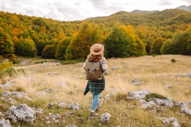 Kayalık bir arazide duran sırt çantalı bir kadın yürüyüşçünün dağ manzarasına bakışı. Şapkalı genç bir kadın yürüyüş ve güneşli havadan hoşlanıyor. Sonbahar, doğa ve özgürlük kavramı.