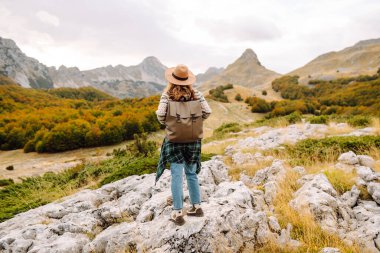 Kayalık bir arazide duran sırt çantalı bir kadın yürüyüşçünün dağ manzarasına bakışı. Şapkalı genç bir kadın yürüyüş ve güneşli havadan hoşlanıyor. Sonbahar, doğa ve özgürlük kavramı.