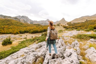 Kayalık bir arazide duran sırt çantalı bir kadın yürüyüşçünün dağ manzarasına bakışı. Şapkalı genç bir kadın yürüyüş ve güneşli havadan hoşlanıyor. Sonbahar, doğa ve özgürlük kavramı.